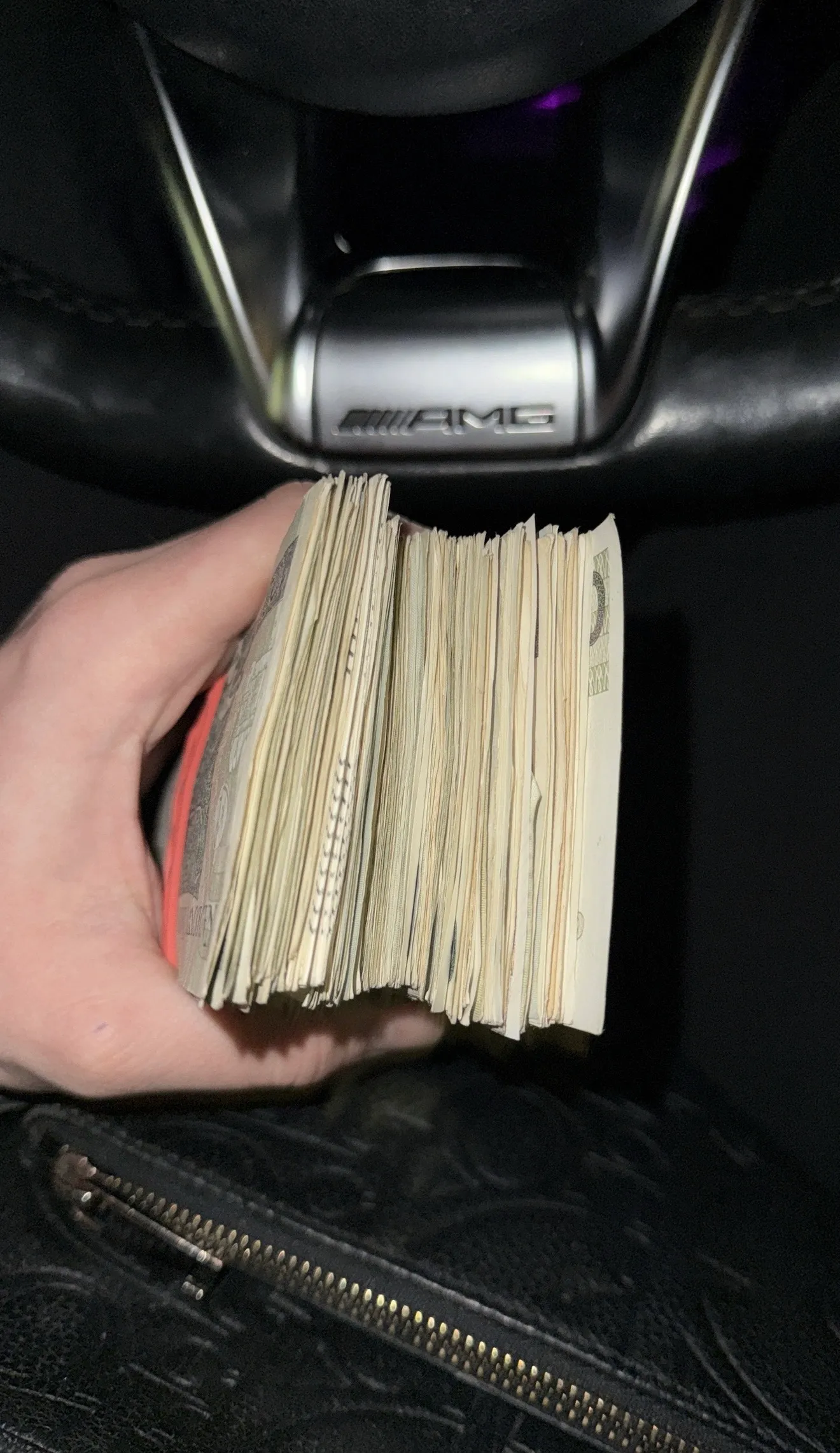 Plik banknotów trzymany w dłoni na tle kierownicy Mercedesa AMG